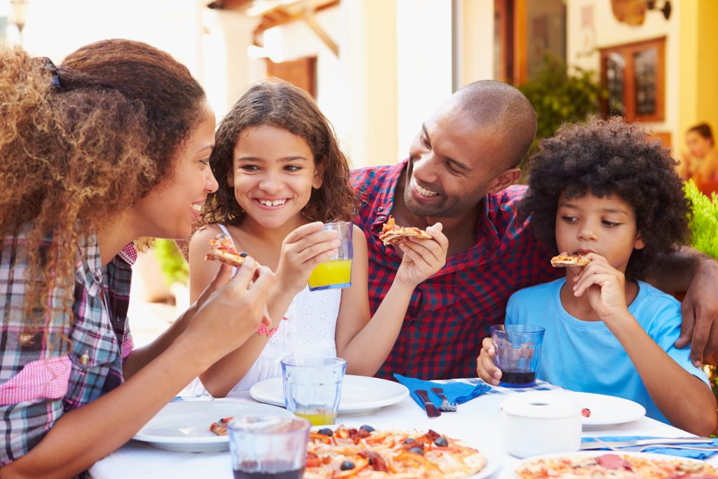 Family Eating Meal At Outdoor Restaurant Together, Smiling, Woman eating a slice of pizzs, gifl drinking orange juice, man smiling at girl, boy eating pizza holding a glass of drink