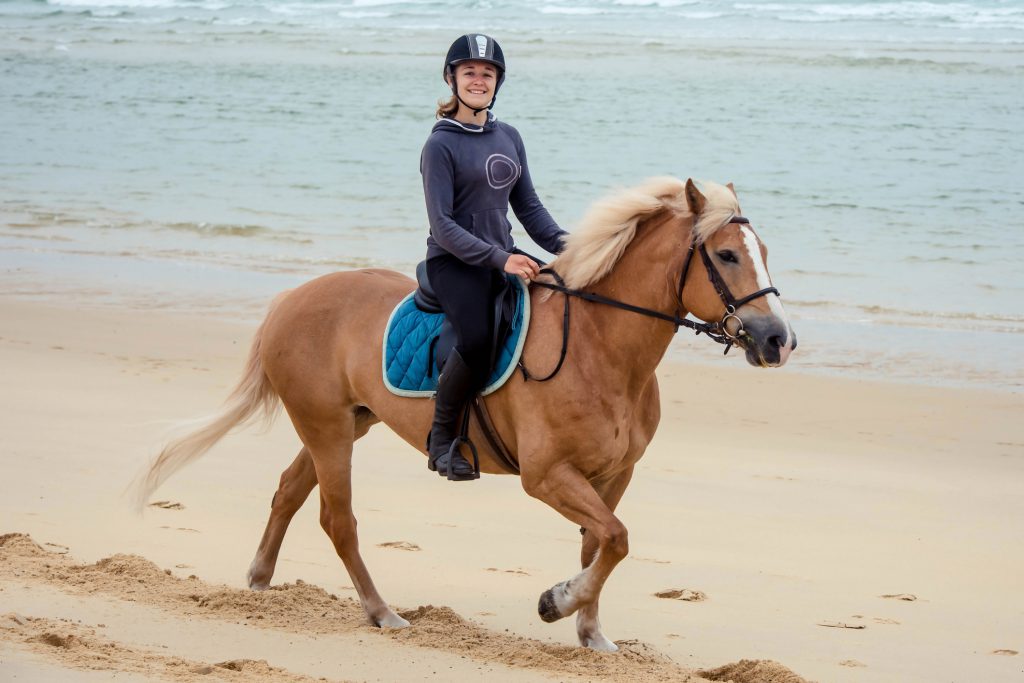 Young Woman riding a brown horse on sand with water in the background. Woman has a helmet on to protect her. 